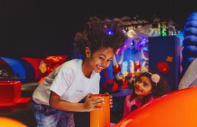 A child with a big smile and wild hair is playing in an indoor playground, leaning over an orange structure.