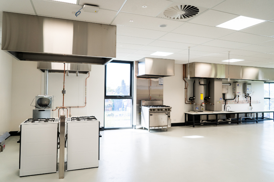 Interior of a modern training commercial kitchen with stainless steel appliances, large hoods, and a view through a window.