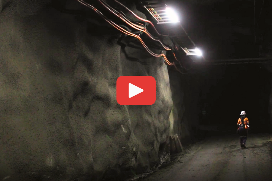 A person in safety gear walks away from the camera in a dimly lit tunnel, with cables running along the tunnel's ceiling.