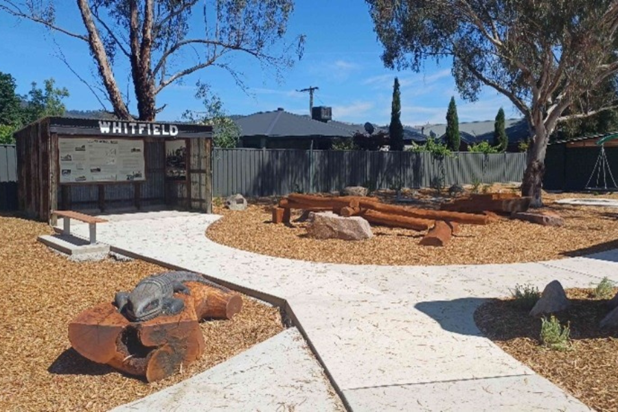 Outdoor adventure playground area in Whitfield featuring curved concrete pathways, wooden benches around a central area, and a large notice board displaying community information.