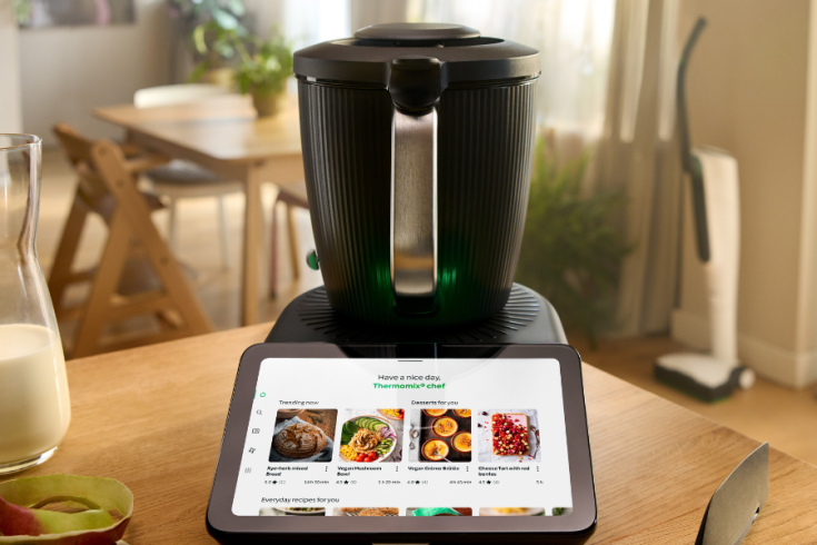 A Thermomix kitchen appliance is on a table next to a tablet displaying recipes. The background shows a chair and part of a dining area.