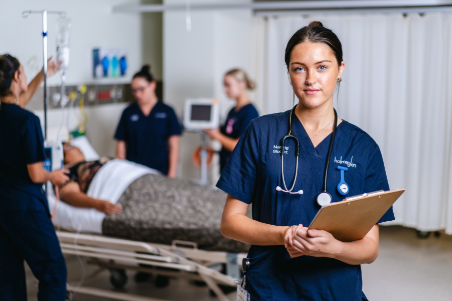 A healthcare professional in navy scrubs holds a clipboard in a medical setting, standing in the foreground. In the background, a patient lies on a hospital bed, attended by team members. The environment includes medical equipment and supplies.