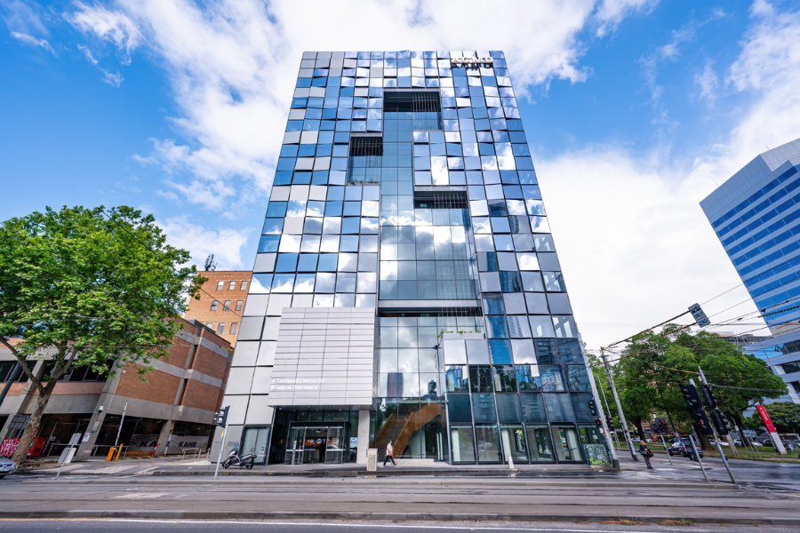 A modern glass building with a facade of reflective squares against a cloudy blue sky. A street with traffic lights and trees is in the foreground. The building is labelled with ACMD.