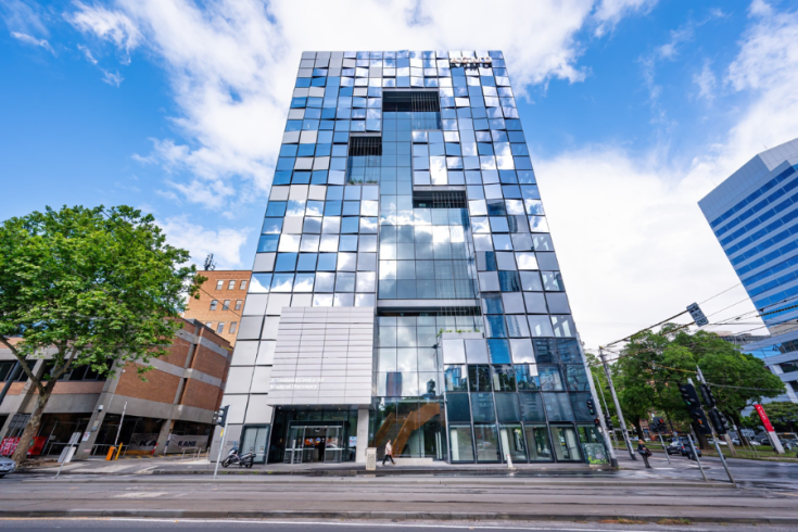 A modern glass building with a facade of reflective squares against a cloudy blue sky. A street with traffic lights and trees is in the foreground. The building is labelled with ACMD.
