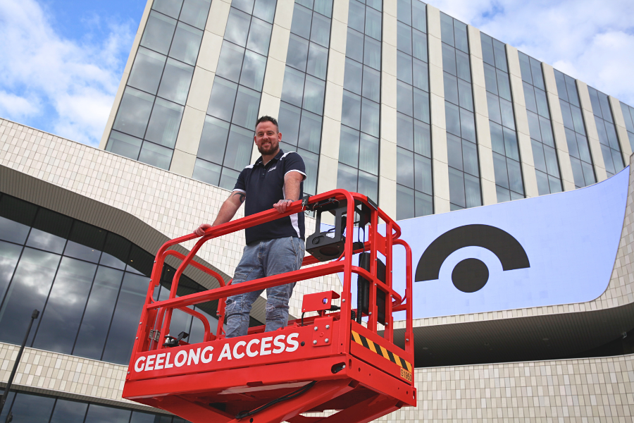 A person in a cherry picker is smiling at the camera. It is outisde a building 