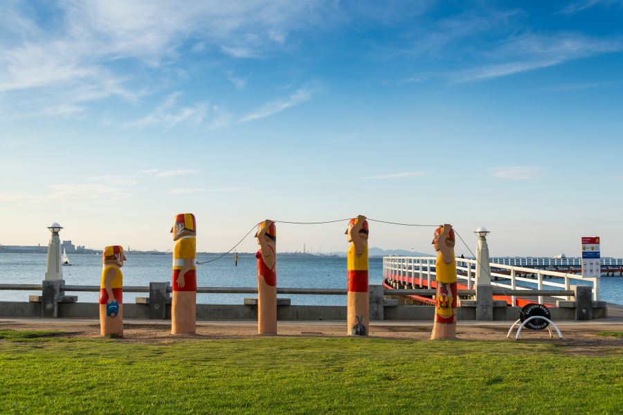 A waterfront view featuring colourful bollards resembling lifeguards in various poses by the Geelong Waterfront. A pier extends into the calm water, with a clear blue sky above.