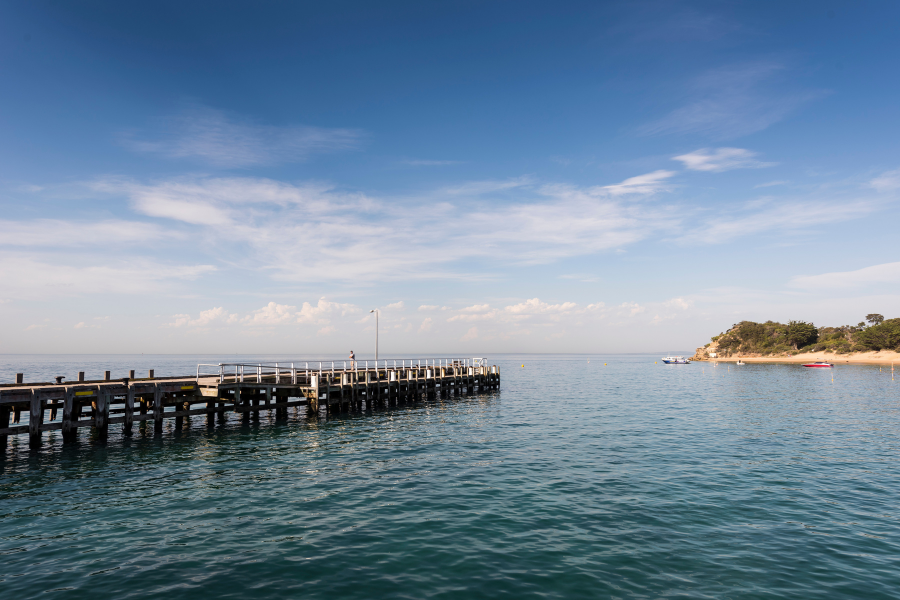 Gold winner of Top Tiny Tourism Town - Portsea A scenic view of a wooden pier extending into the calm, blue waters of Portsea Pier. The sky is clear with a few clouds, and a small peninsula with trees is visible in the distance.