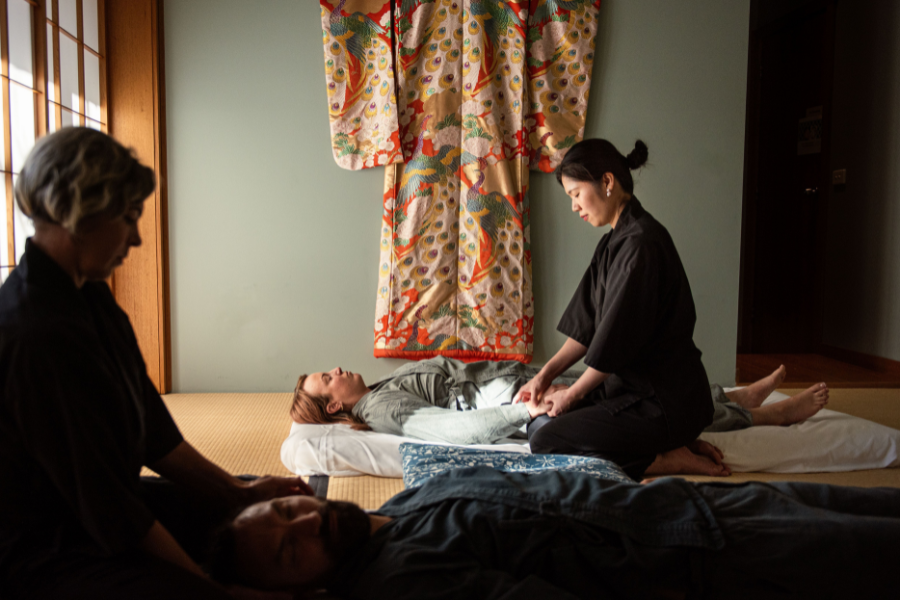 Two people receive a relaxing massage in a tranquil room. A colourful traditional kimono hangs on the wall. The space is softly lit, creating a serene atmosphere at the Shizuka Ryokan. 