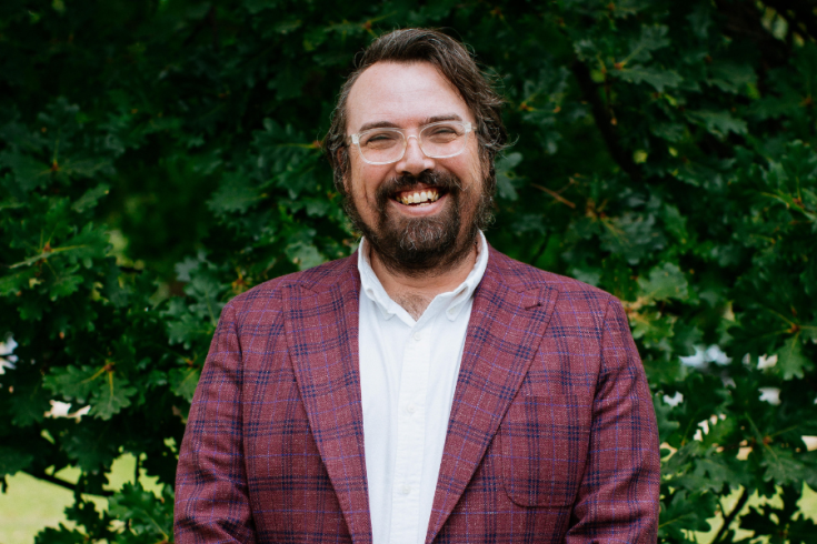 Greg Jennings smiling standing in front of a green leafy wall in a suit with purple plaid jacket