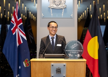 Dr Rongbin Xu standing at a podium smiling, with an "Excellence Award 2023-24" plaque visible. Behind are the flags of Australia and the Australian Aboriginal flag.