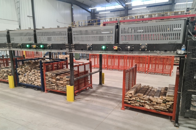 Interior of a warehouse with machinery for processing lumber, featuring stacks of wood on pallets and industrial equipment with green status lights on.