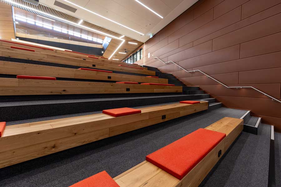 Interior of a modern lecture hall featuring tiered wooden seating with red cushions and stair access on the side.