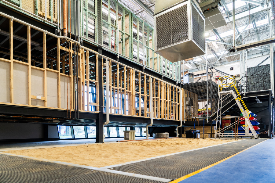 Interior view of a large industrial site featuring exposed wooden framing and mechanical systems, with a sandpit in foreground and a mobile stairway in background.