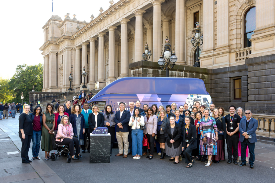 A group of diverse individuals posing in front of the Parliament House of Victoria, Australia, with a large blue small business bus behind them.