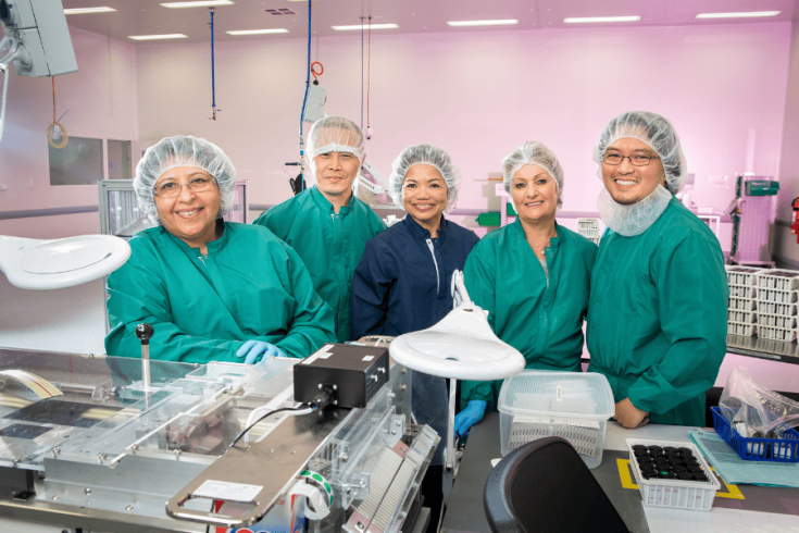 5 smiling medical staff in scrubs and hairnets standing behind laboratory equipment.