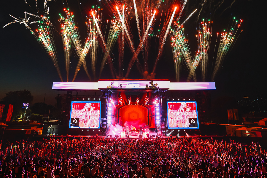 A large crowd attends an outdoor concert at night to celebrate the end of the Formula 1 Australian Grand Prix. The main stage displays bright lights and a large screen, while vibrant fireworks in various colours explode in the sky above.