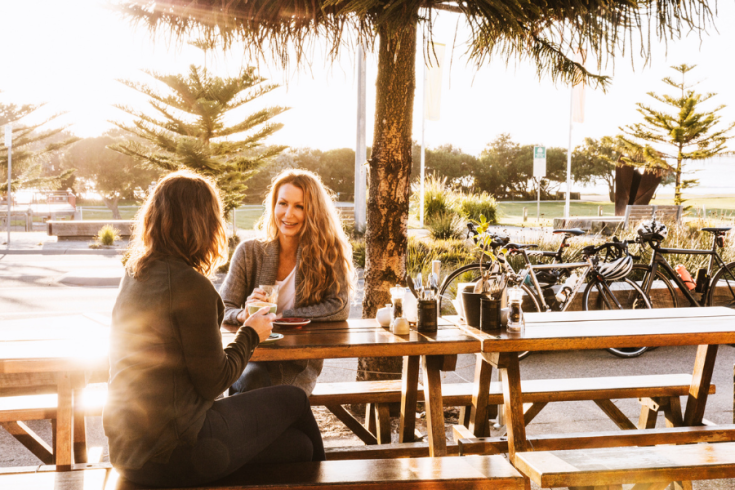 Two people seated at a wooden outdoor table at Moons Cafe in Lorne, with bicycles, palm trees and a sunlit foreshore in the background.