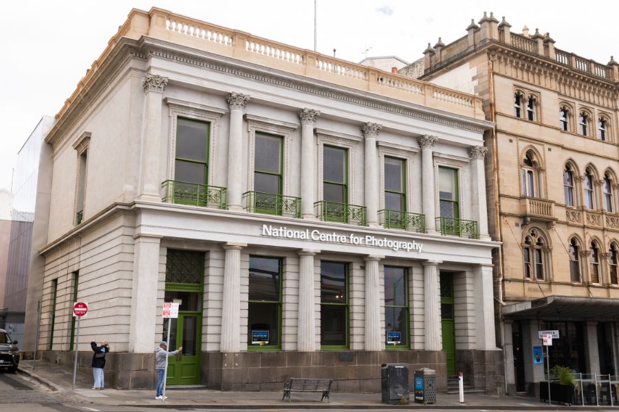 A historic building with stone columns and large windows, labelled: National Centre of Photography. It has a green door and green window frames. It's next to another historic building.