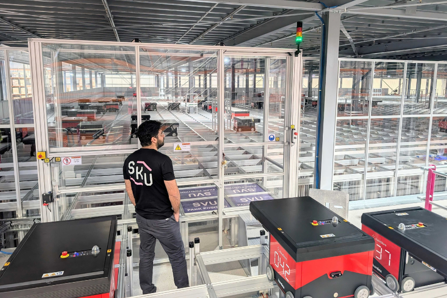 A person wearing a black SKU t-shirt stands in a high-tech warehouse. They are observing an automated system with red and black robots moving along tracks. The environment includes metal structures and large glass panels.