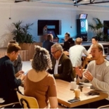A group of people are gathering in a co-working space, participating in a networking event. There are television screens on the walls, as well as indoor plants in the room. 