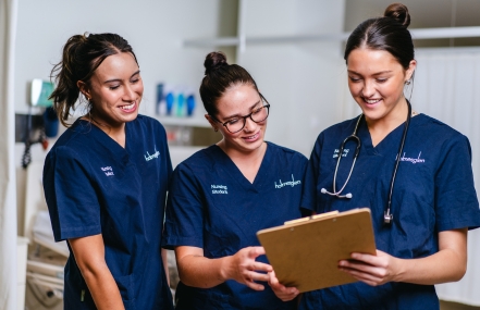 Three healthcare professionals in scrubs, two smiling and one holding a clipboard, are having a discussion.