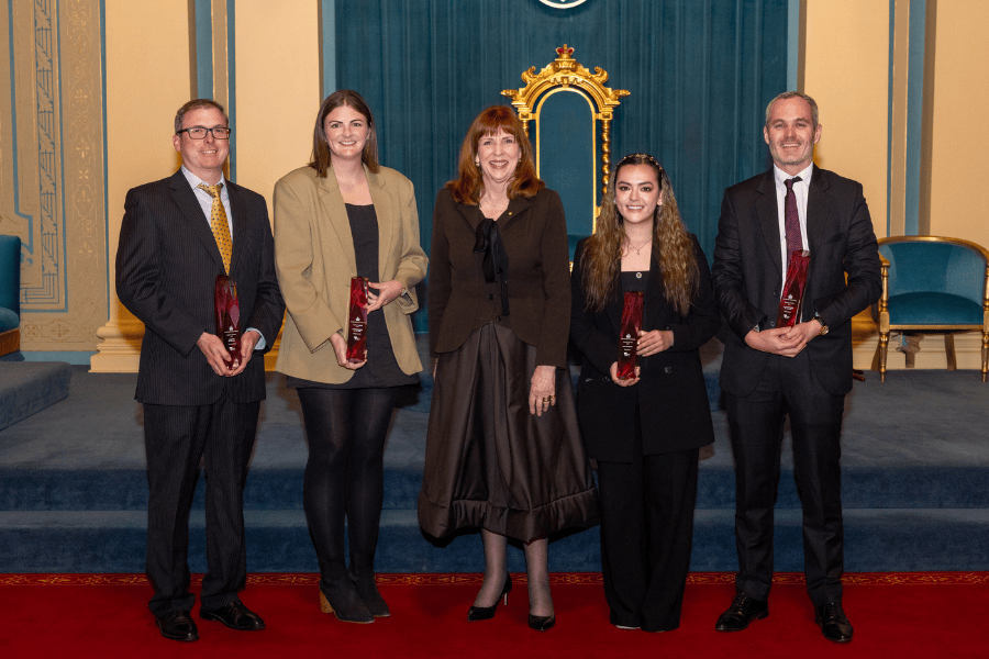 5 people standing in a row all smiling, 4 are holding up awards