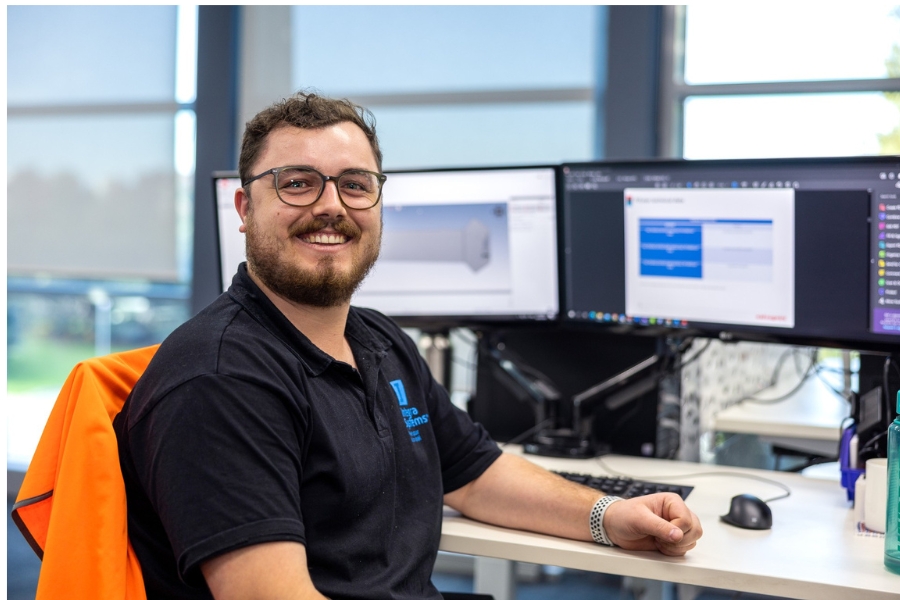 A man with a beard and glasses is smiling. He is sitting in front of two computer screens