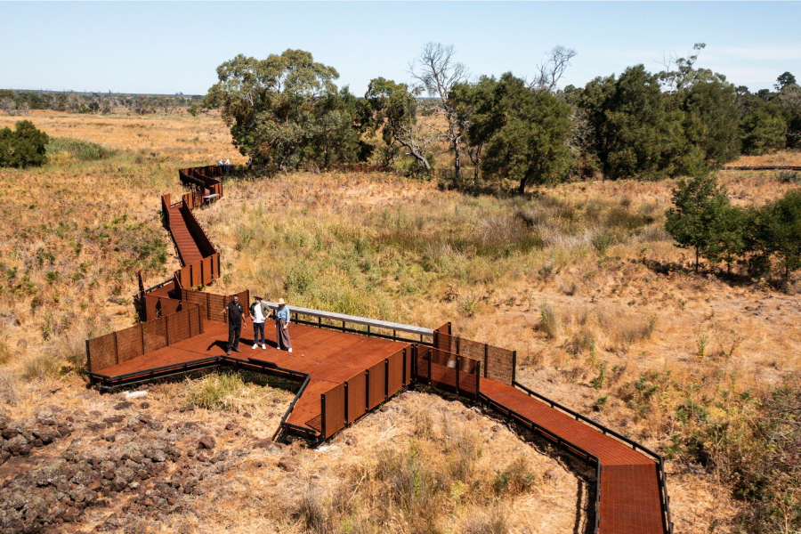 Aerial view of landscape and walkways and viewing platform at Budj Bim. There are 3 people on the platform and one is gesturing to the landscape. 