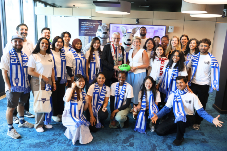 A group of people, including 2 mascots, pose for a photo while holding a AFL football. Many in the group are wearing white t-shirts and blue and white striped scarves (North Melbourne Kangaroos).