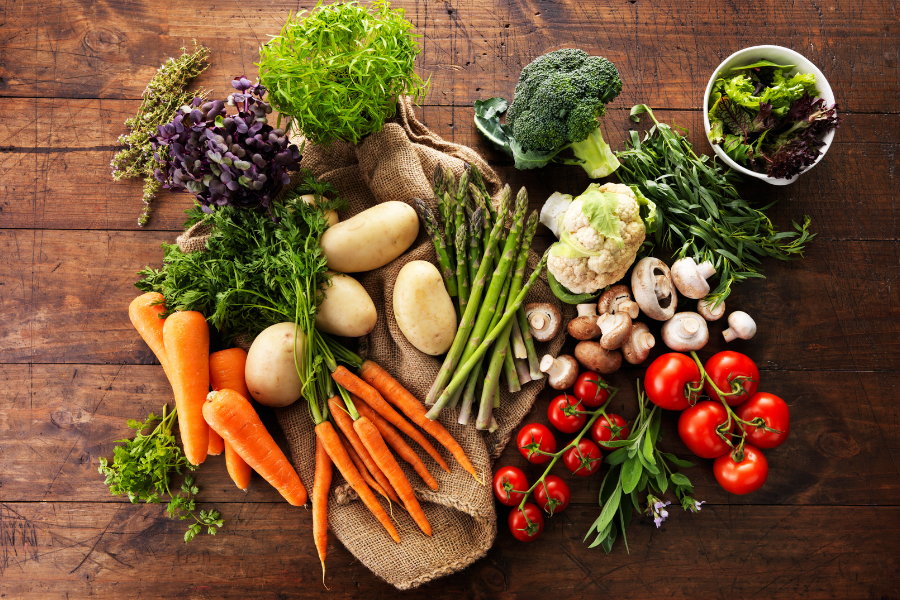 A variety of fresh vegetables arranged on a wooden surface, including carrots, potatoes, asparagus, broccoli, cauliflower, mushrooms, cherry tomatoes, and leafy greens.