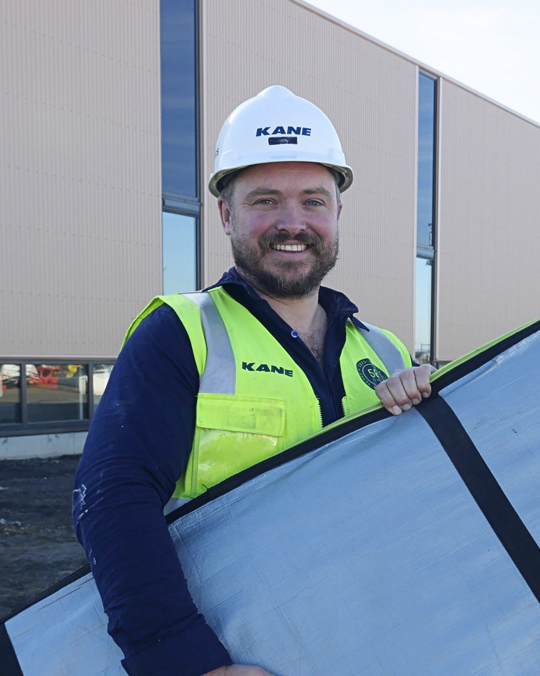 Man wearing a hard hat and hi vis vest is smiling and carrying a surfboard. he's standing in front of a construction site