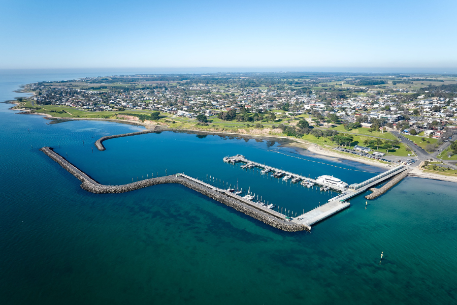 Gold winner of Top Small Tourism Town - Portarlington Aerial view of Port Fairy in Victoria, Australia, showcasing a marina with multiple boats docked along piers. The town is visible in the background with a coastline stretching out under a clear blue sky.
