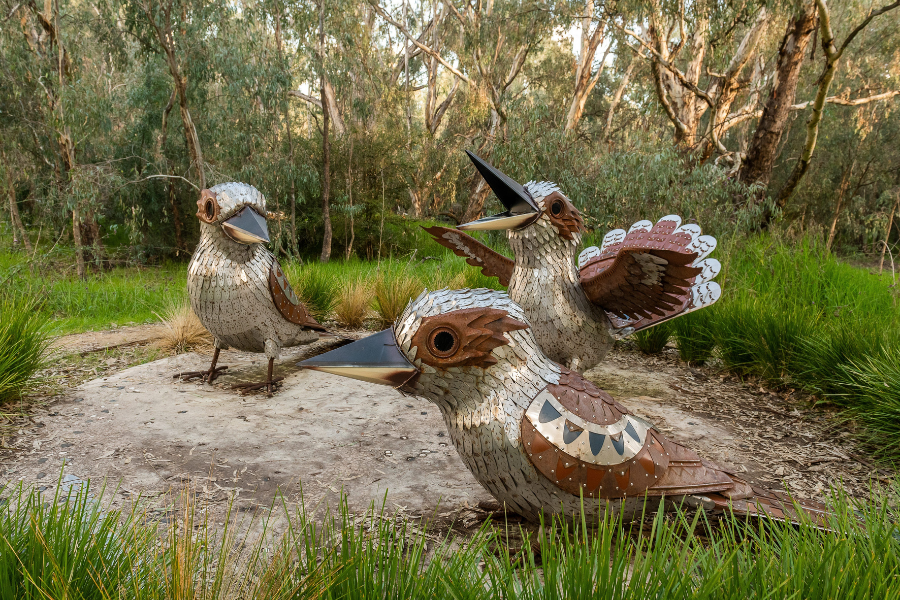 Three metallic sculptures of kookaburras are displayed outdoors among Australian bushland