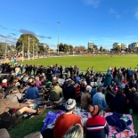 View from behind of spectators sitting on the ground looking towards grassy oval on a sunny day with the Melbourne CBD in the background