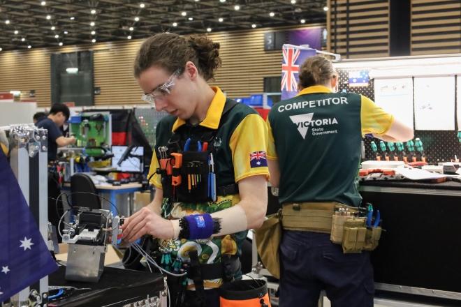 A competitor in a yellow and green uniform labeled  works diligently on machinery at a workbench