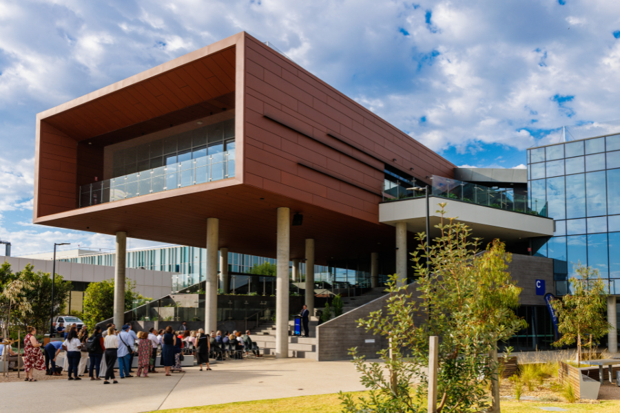 Exterior view of the learning hub with a prominent overhanging structure, wide staircases, and surrounding outdoor seating areas. There is a group of people gathered around the entrance. Lush greenery and a partly cloudy sky can be seen in the background.