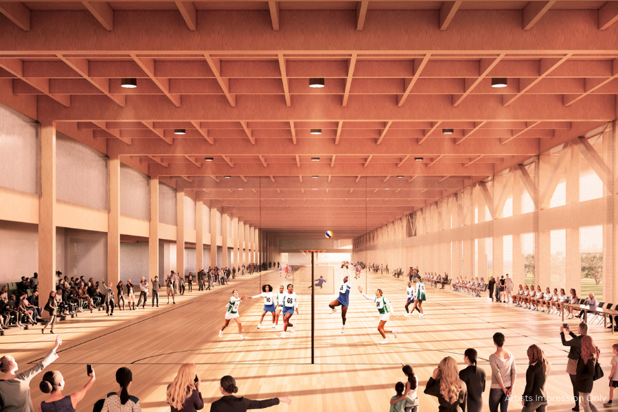 Interior of a spacious modern building with a Netball game in progress. Spectators are seated on either side of the court, watching intently. The architecture features wooden beams and large windows providing ample natural light. Artists impression only. 