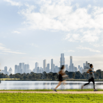 Three individuals running along a lakeside path with the Melbourne skyline in the background.
