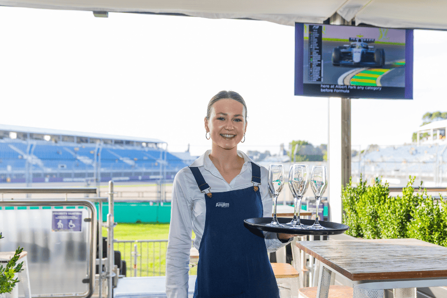 Claire Valentine in a blue apron holding a tray with wine glasses, standing at a bar with a racetrack in the background and a television screen displaying a Formula 1 race.