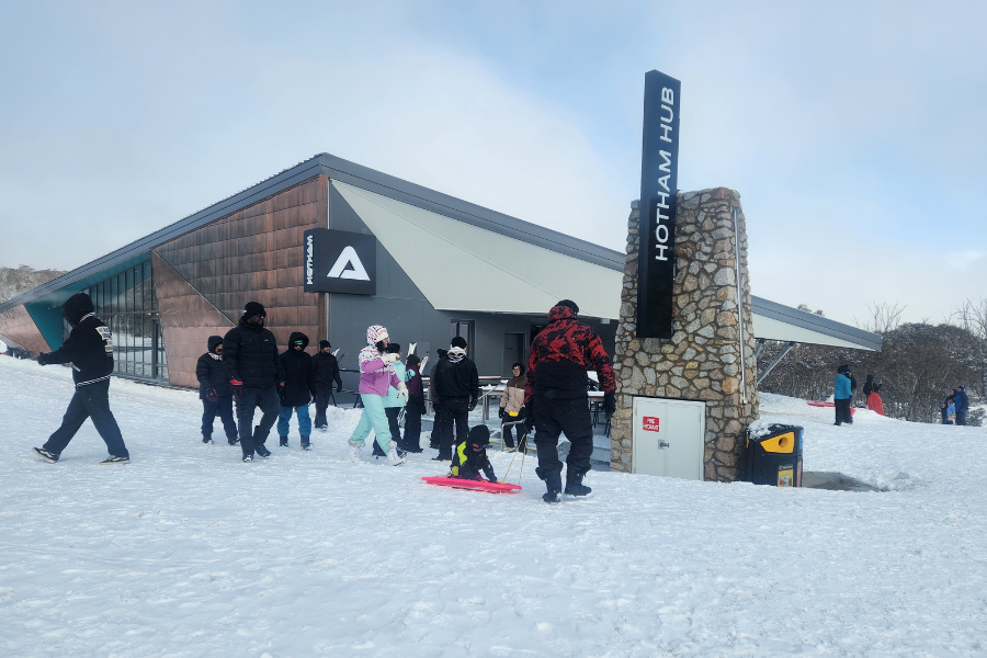 Group of people outside Hotham Hub at a snowy location, some carrying snowboards.