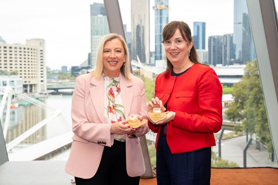Chief Executive Officer of Melbourne Convention Bureau, Julia Swanson and Chief Executive Victorian Convention and Event Trust, Natalie O’Brien AM hold a slice of cake each infront of a window with Melbourne city skyscrapers in the background.