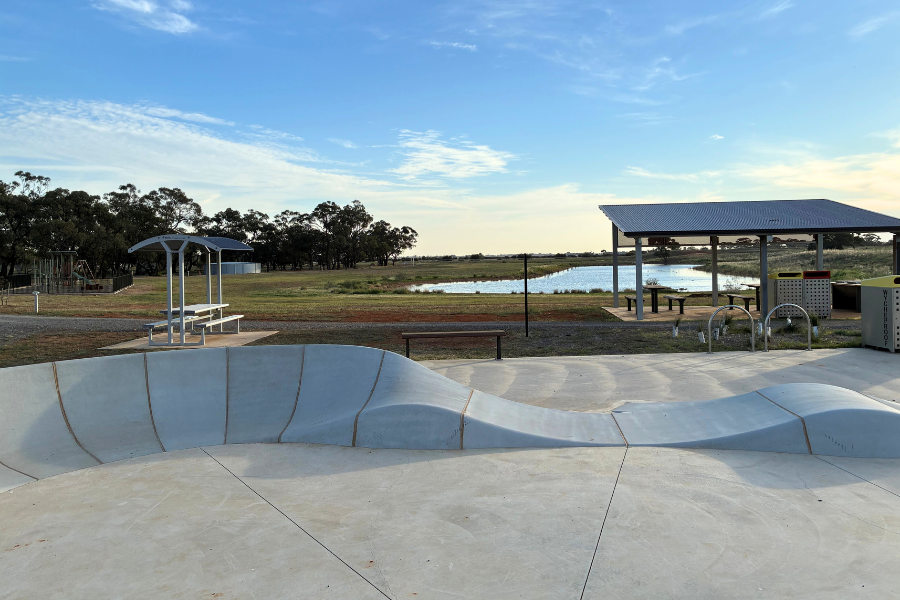 A skate park in Wycheproof with smooth, curving concrete ramps is set under a clear blue sky. Nearby, there's a covered picnic area with benches. In the background, a grassy field stretches towards a body of water, bordered by trees.