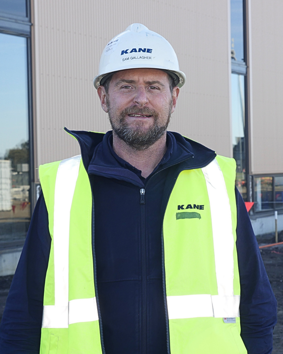 Close up of a man with a beard wearing a hard hat, standing in front of a construction site