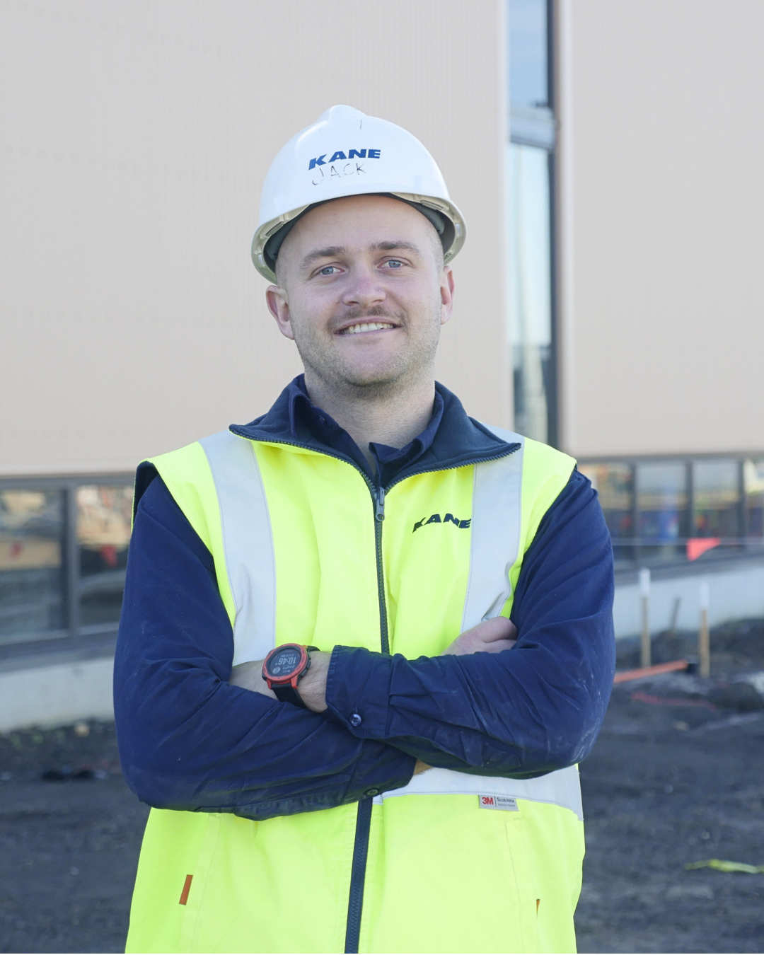 A man is smiling at the camera with his arms crossed. He's wearing a hard hat and hi vis and is standing in front of a construction site