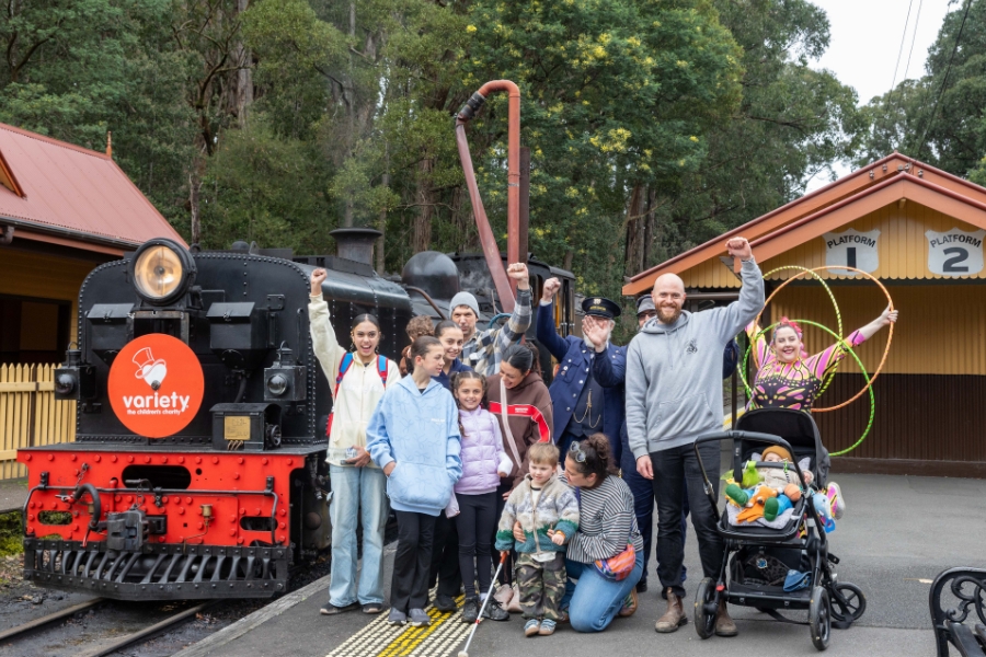 A group of people pose with a fist raised in front of a vintage steam train on a station platform. The front of the train displays the Variety logo. A person in a conductor's uniform stands in the group. One of the people is holding colourful hula hoops and a child sits in a stroller with toys. 