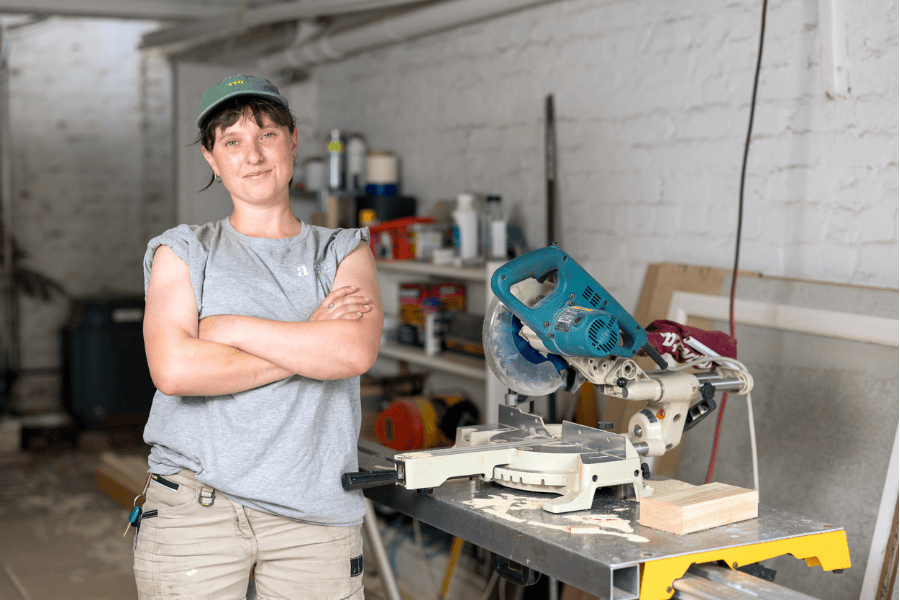 Natasha Eastman-Nagle wearing a cap and a gray t-shirt stands with crossed arms in a workshop, smiling slightly at the camera. Behind them, a workbench holds various woodworking tools including a saw.