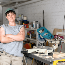 Natasha Eastman-Nagle wearing a cap and a gray t-shirt stands with crossed arms in a workshop, smiling slightly at the camera. Behind them, a workbench holds various woodworking tools including a saw.