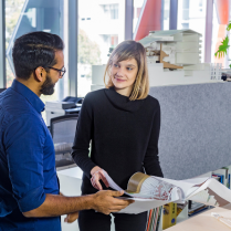 Two professionals discussing over a book in a bright office space, surrounded by architectural models and plants.