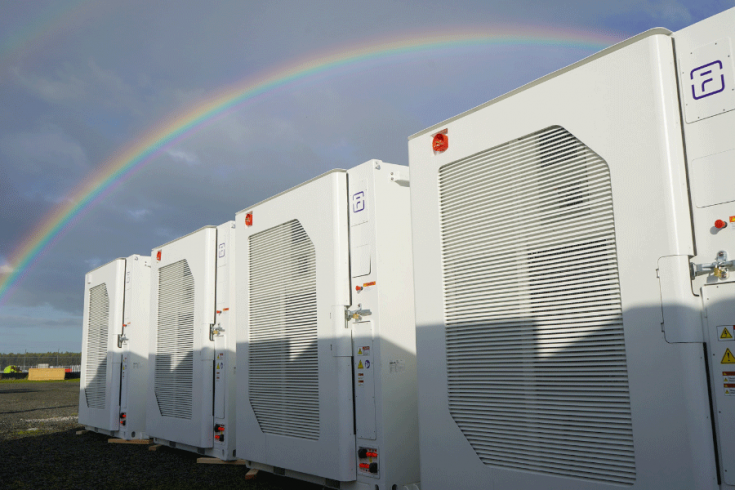 Row of large industrial battery storage under a bright sky with a vivid rainbow arching in the background.