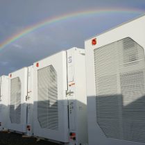 Row of large industrial battery storage under a bright sky with a vivid rainbow arching in the background.